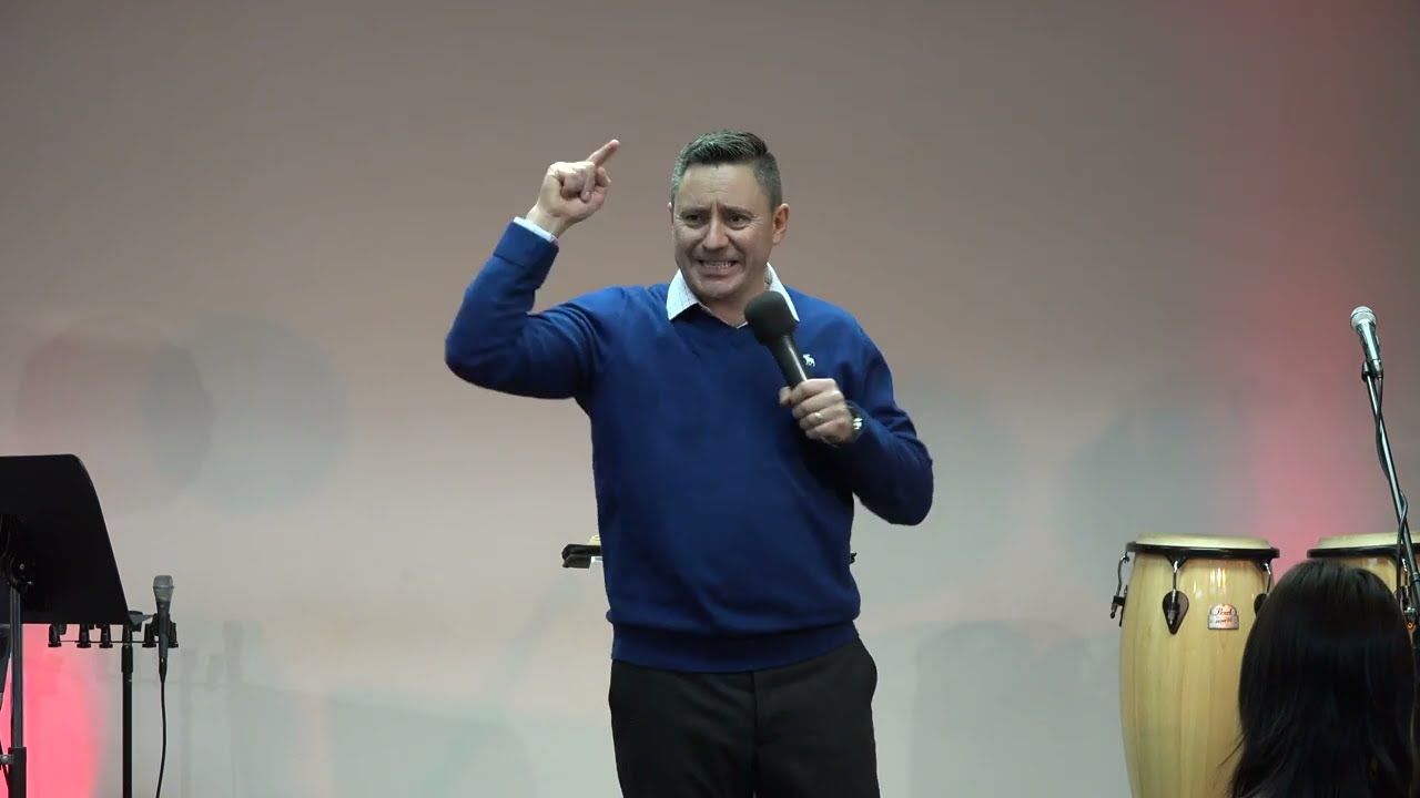 Ruslan Murin in a blue sweater preaching into a microphone and pointing upward.