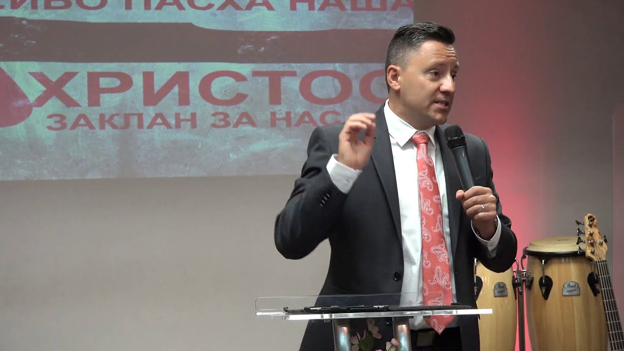 Ruslan Murin in a dark suit and tie preaching into a microphone in front of a projected slide.
