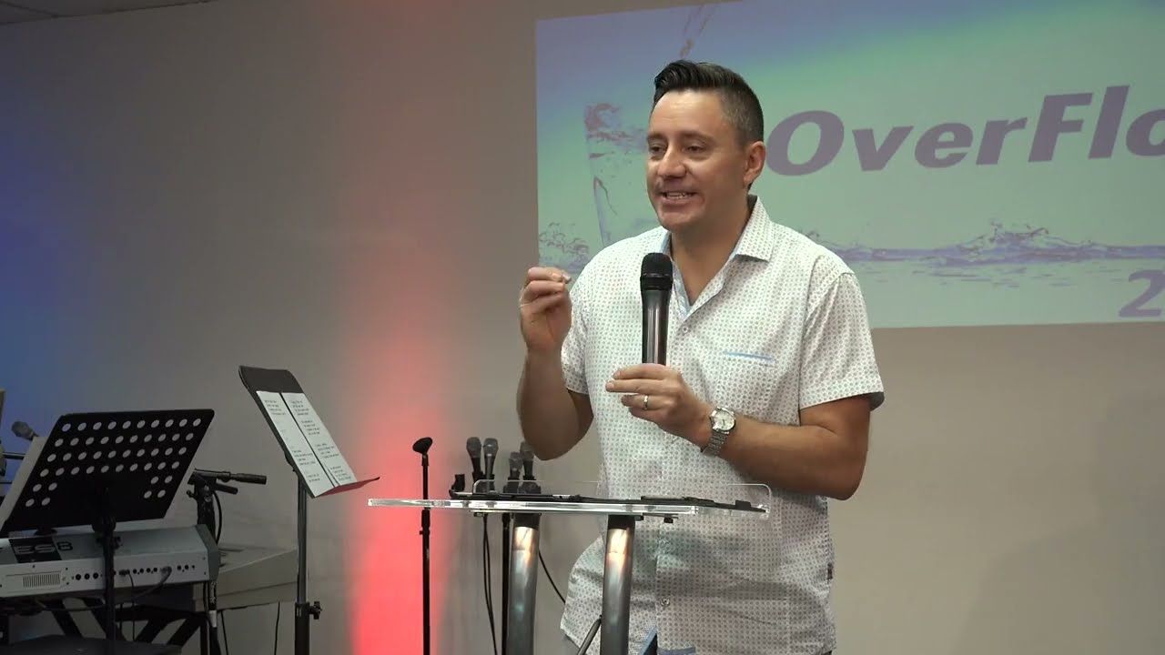 Ruslan Murin preaching into a microphone at a clear lectern and gesturing with one hand.
