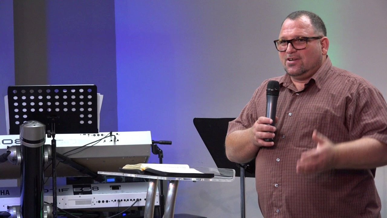 A man in glasses preaching into a microphone and gesturing with one hand.