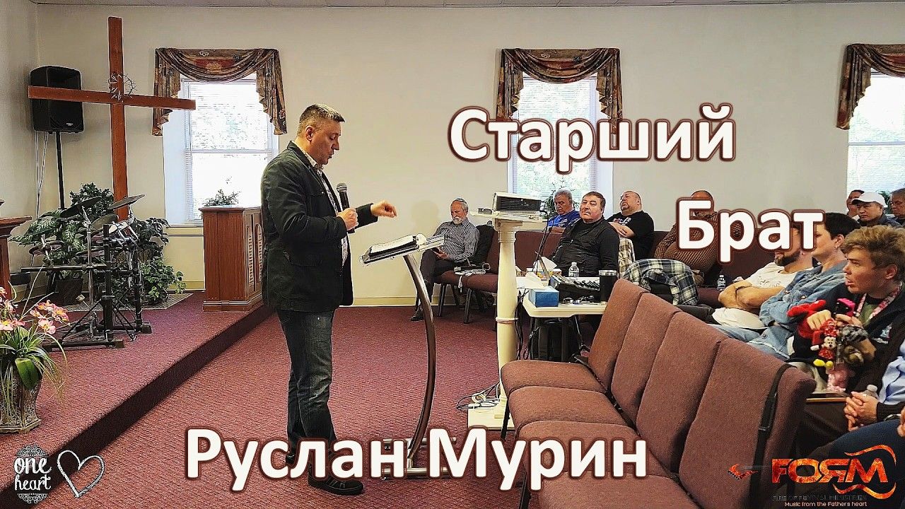 Ruslan Murin preaching with a microphone beside a lectern, facing people seated in rows.